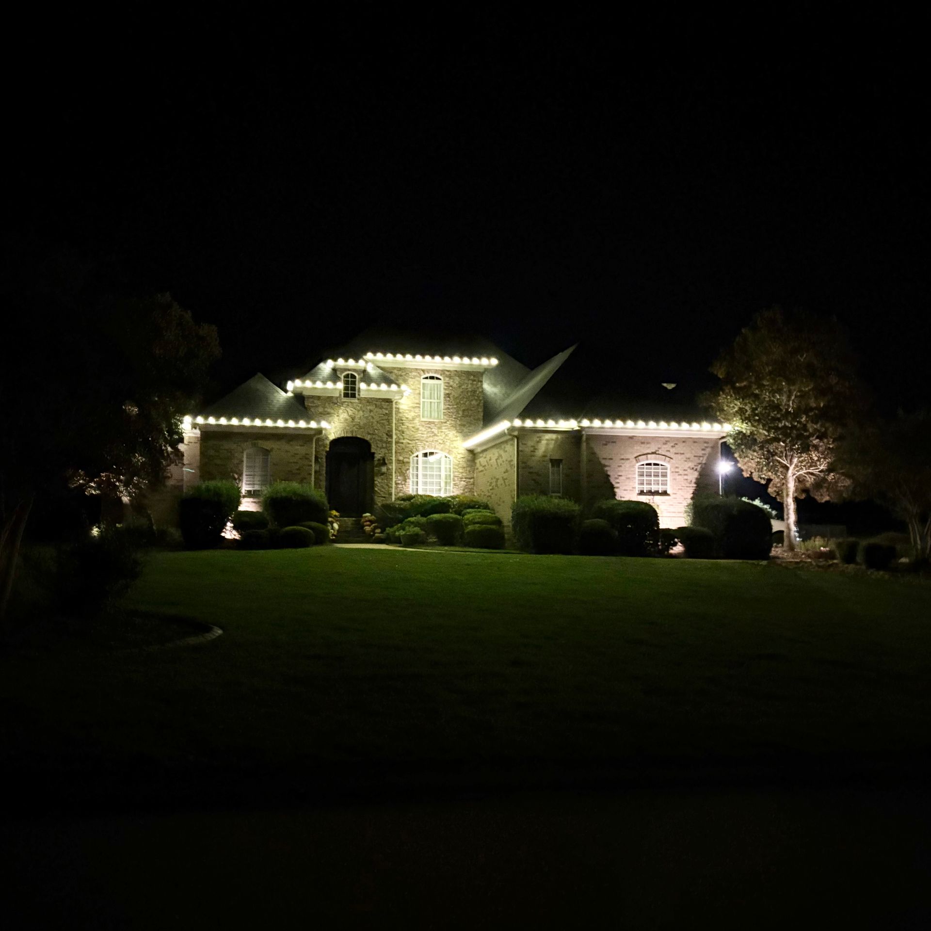 Nighttime view of a stone house lit by white string lights, with a dark lawn and trees in front