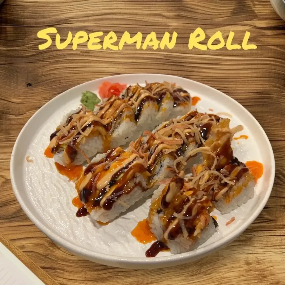 Plate of sushi rolls drizzled with sauces on a wooden table, labeled “Superman Roll”