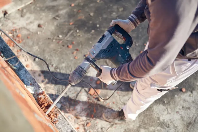 Person using a jackhammer on a brick surface, wearing gloves and light pants.
