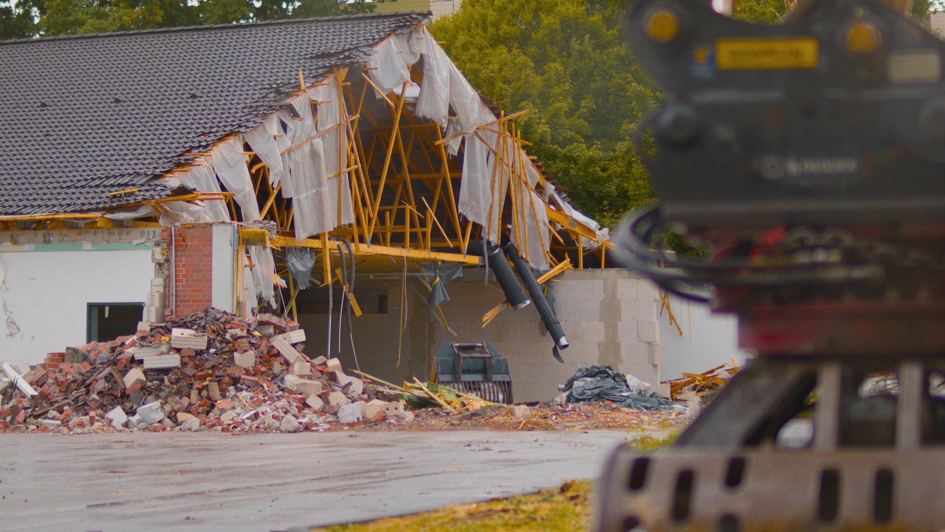 Building being demolished, partially collapsed roof, red brick debris, excavator arm in foreground.