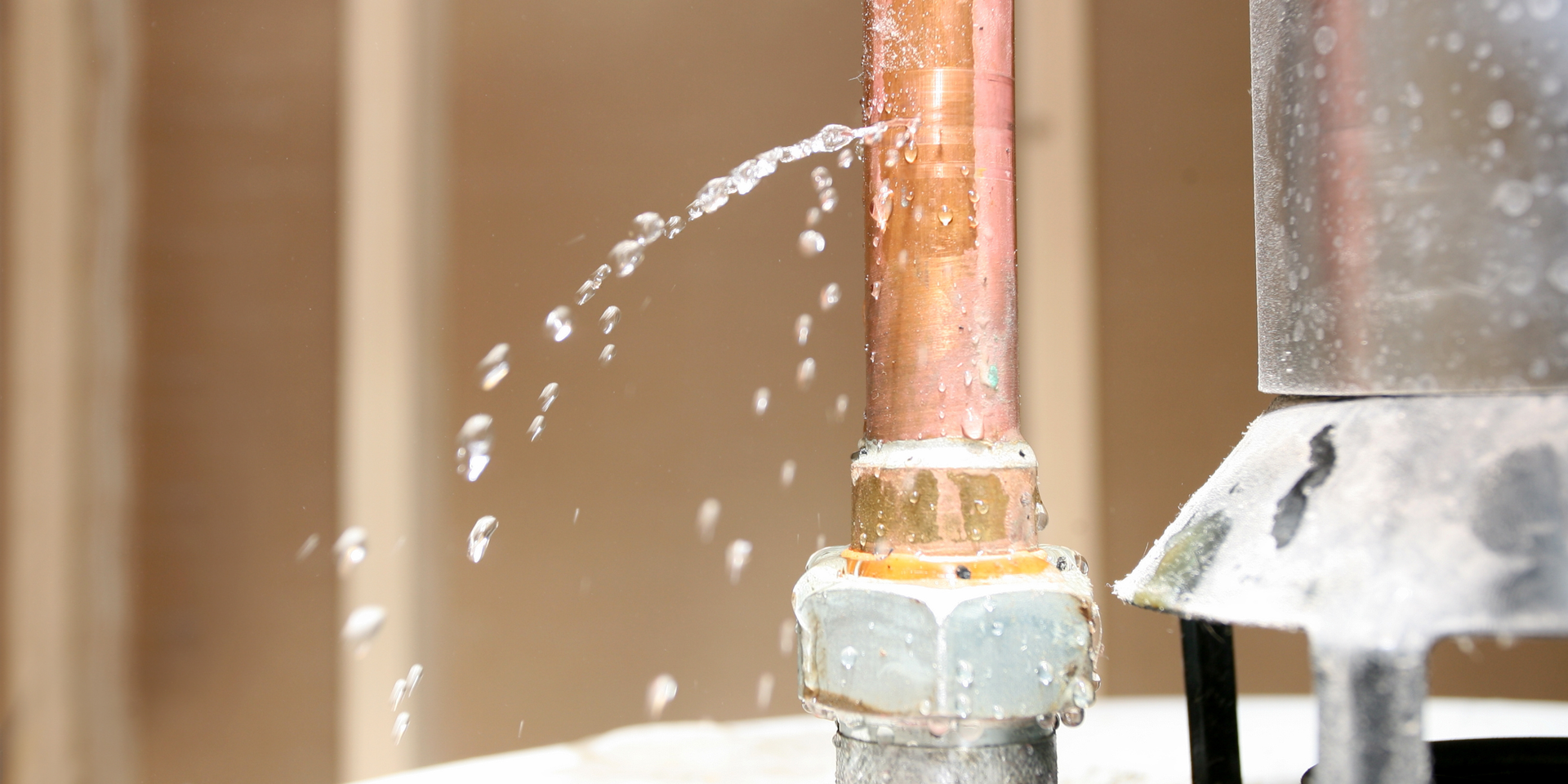 A copper pipe leaks water, spraying droplets. The pipe is connected with a metal fitting against a blurred wooden wall.