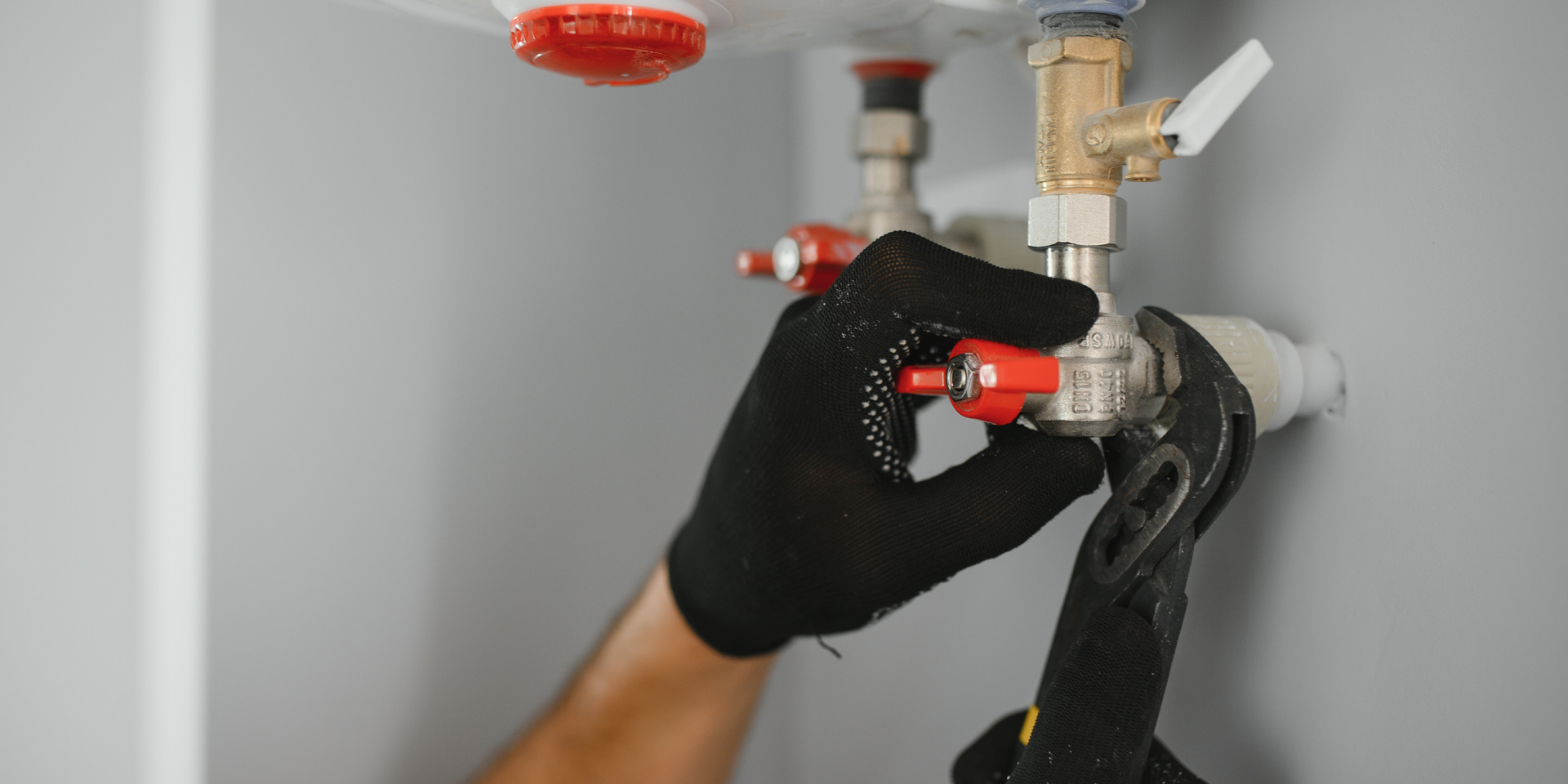 Plumber using pliers on pipes, wearing black gloves. Setting is a wall with plumbing.