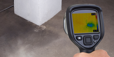 Hand holding thermal camera showing blue spot on floor near white structure.