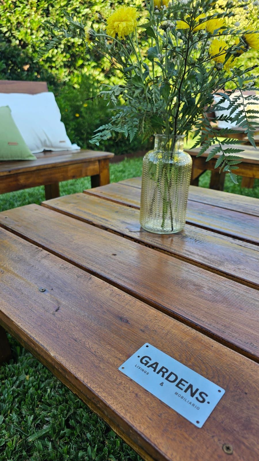Zona de estar al aire libre con muebles de madera, cojines blancos y luces de cadena, ubicada en un patio con césped.