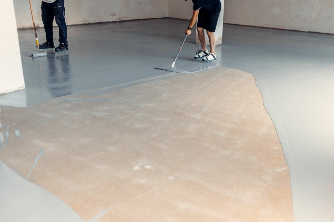 Two workers spreading a light tan coating on a concrete floor indoors