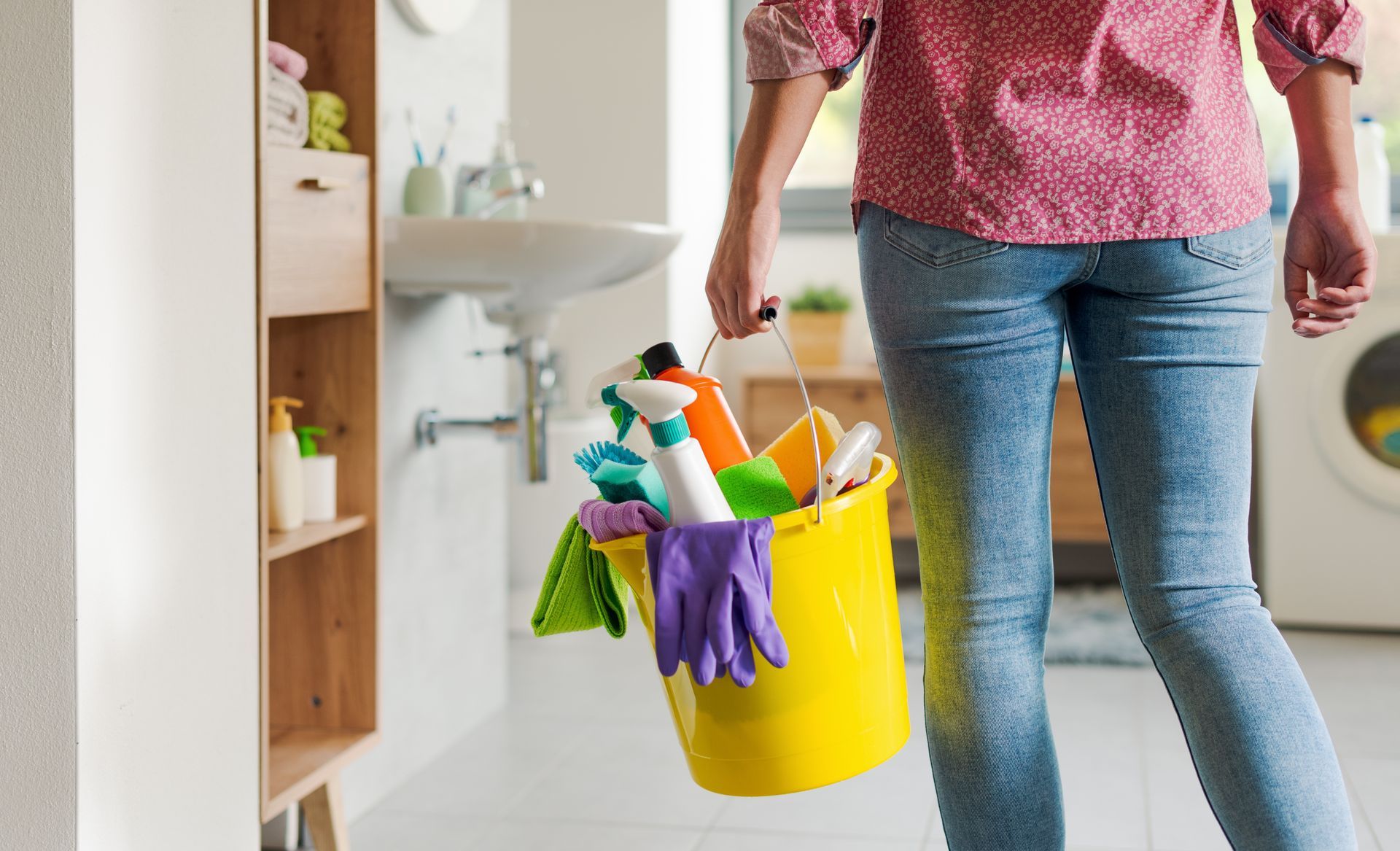 A woman is carrying a yellow bucket filled with cleaning supplies.