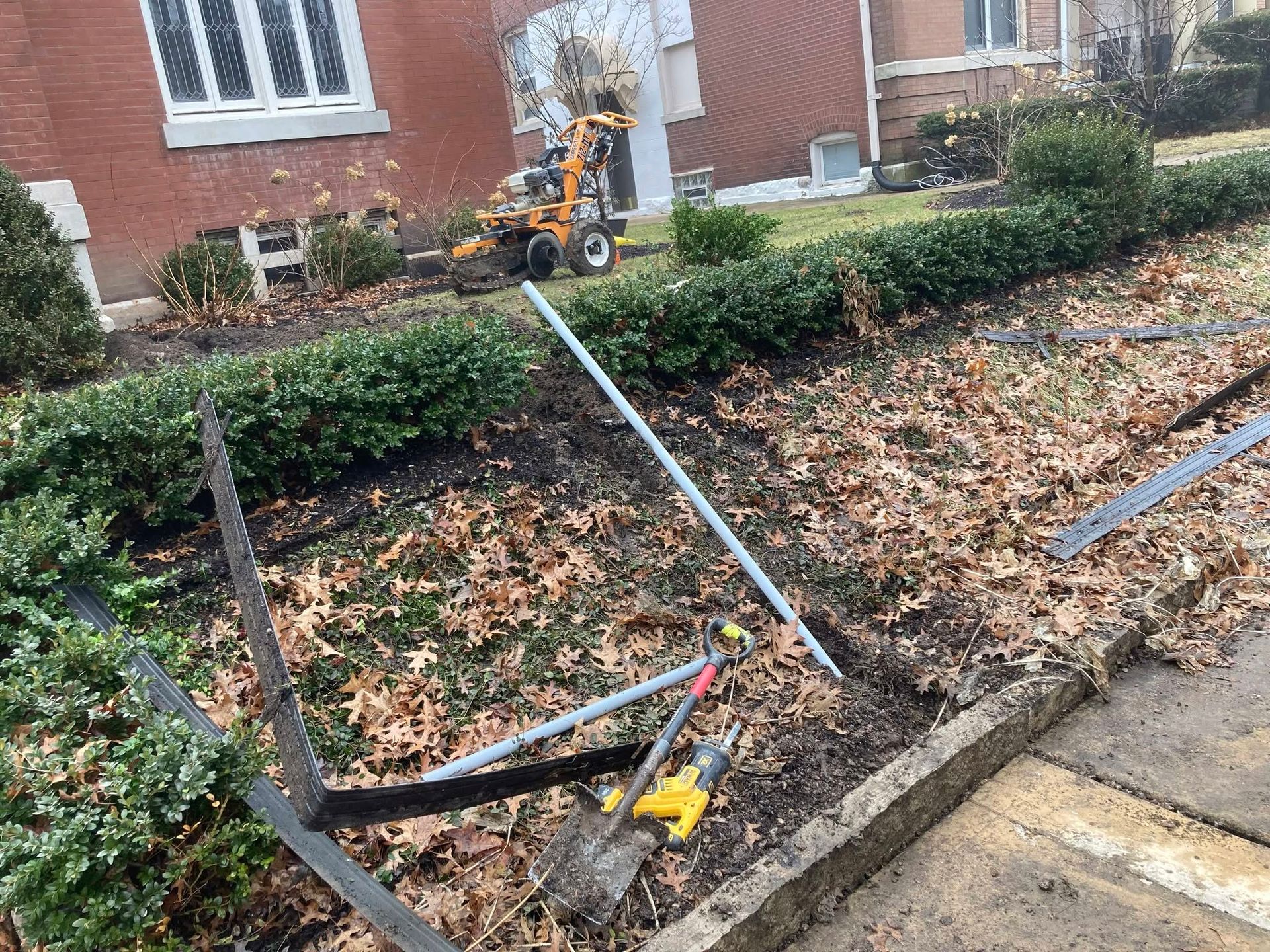 Broken garden fence with fallen metal rails, hedge, and a yellow power tool near a brick house.