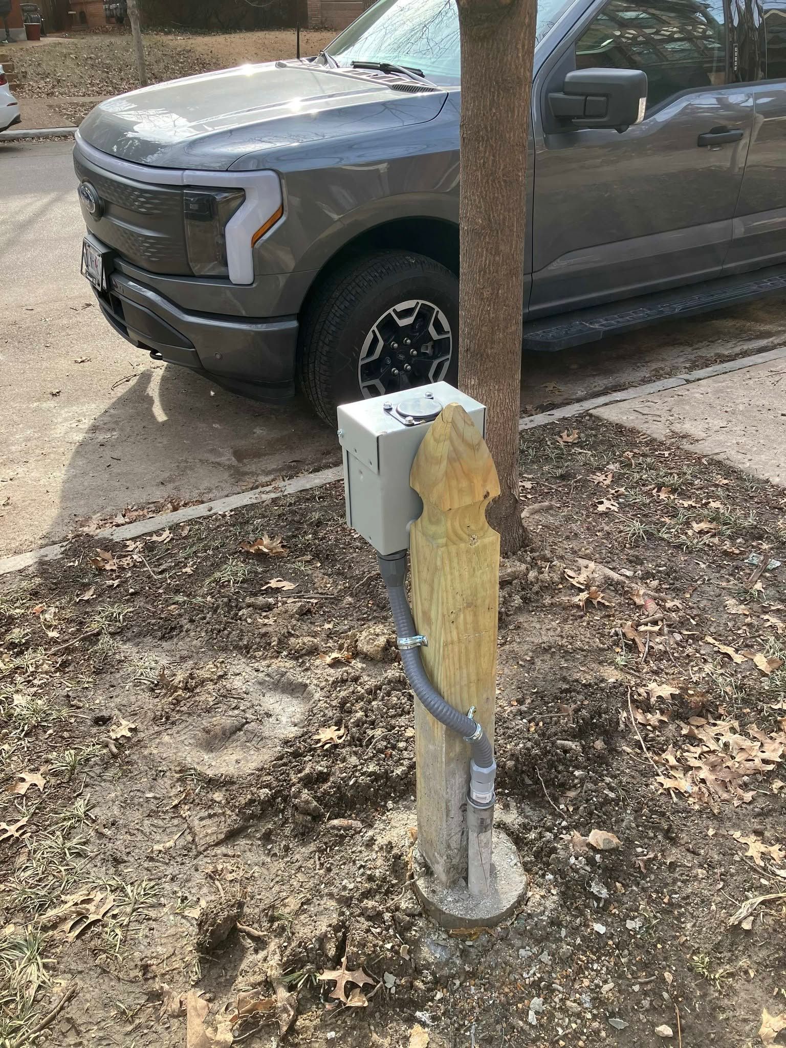 Gray pickup truck parked beside a utility post with a yellow meter box in a wooded yard