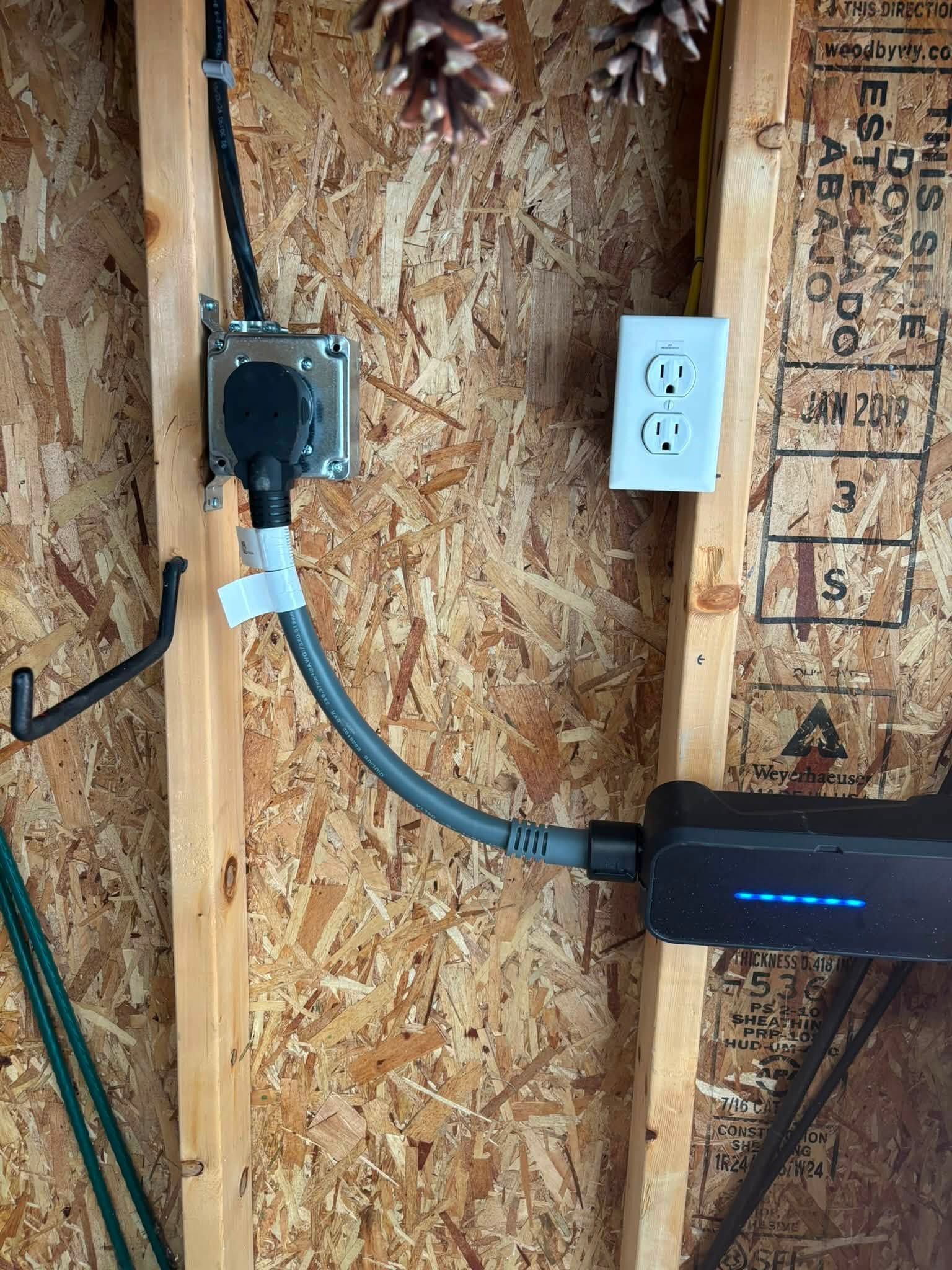 Wall-mounted outlet and metal box with a blue cable connected to a black device on plywood sheathing.