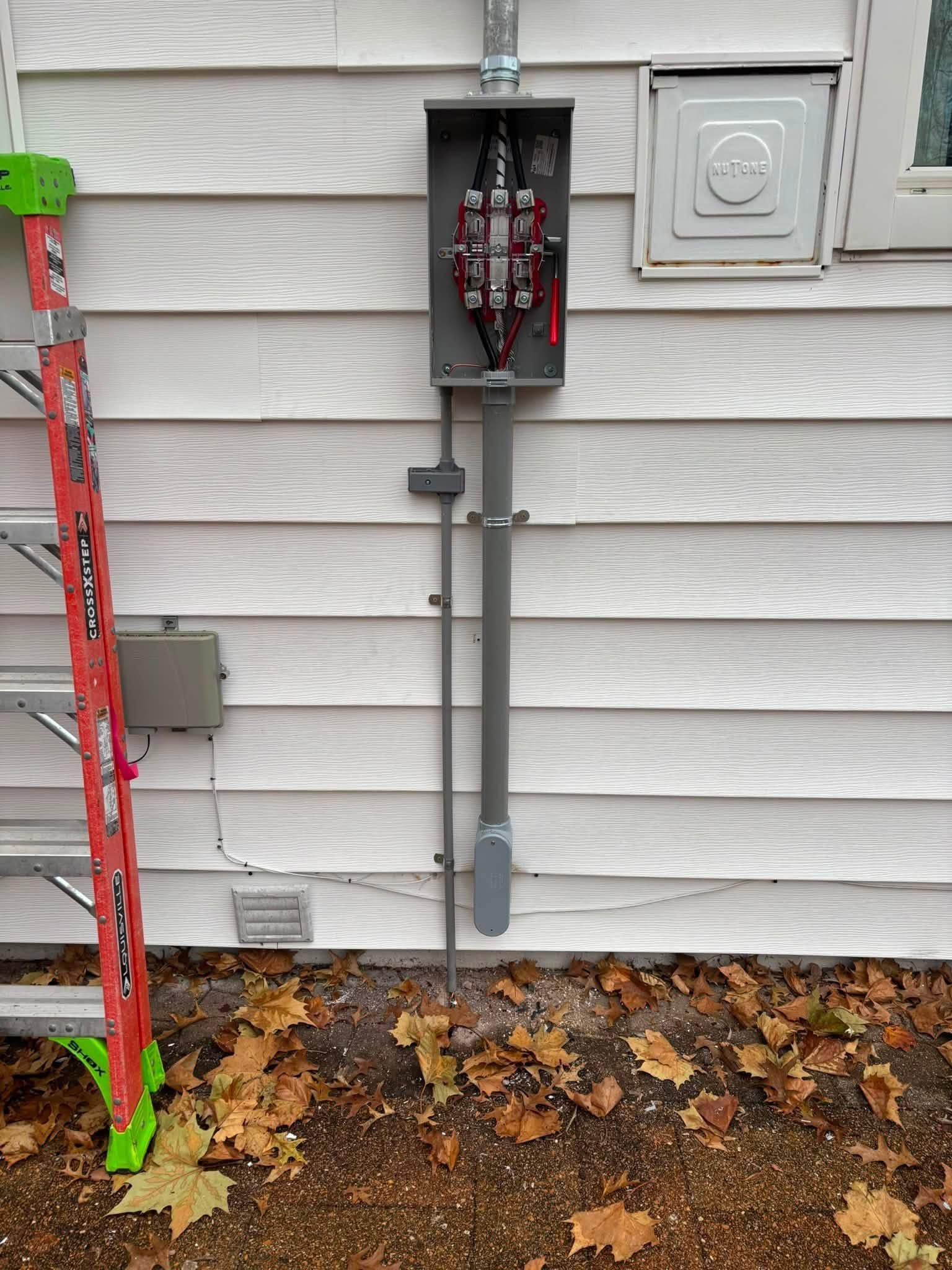 Exterior wall electrical panel with exposed wiring and conduit beside a red ladder on the ground.