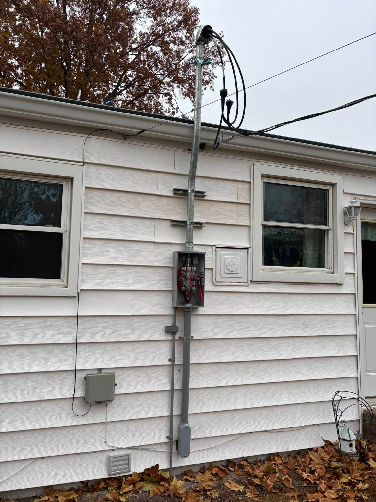 Side of white house with electric meter box, conduit, and overhead service lines outdoors.