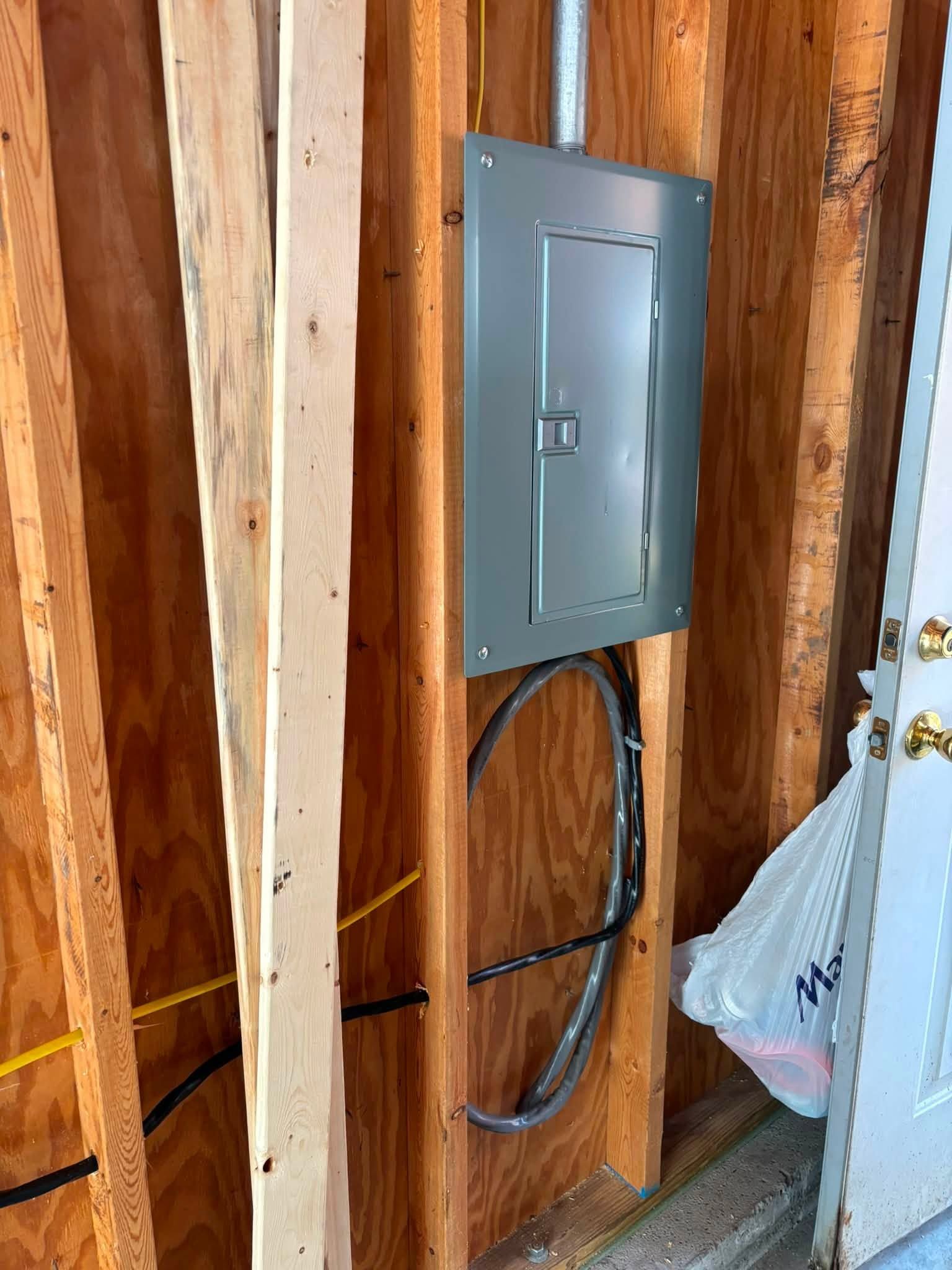 Electrical breaker panel mounted on unfinished wooden studs, with exposed wiring and a plastic bag nearby