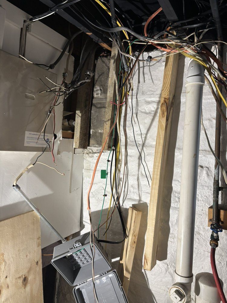 Cluttered basement utility area with exposed wires, hanging cables, and a wall-mounted electrical panel.