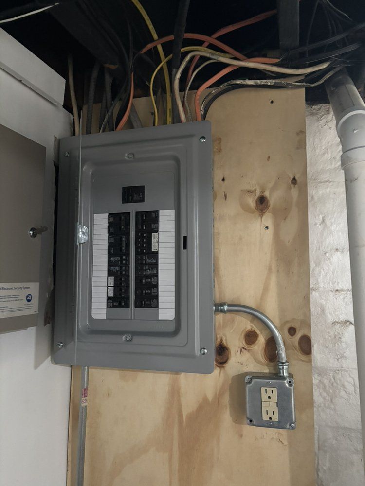 Electrical breaker panel mounted on a wall with exposed wiring and a nearby outlet in a utility space
