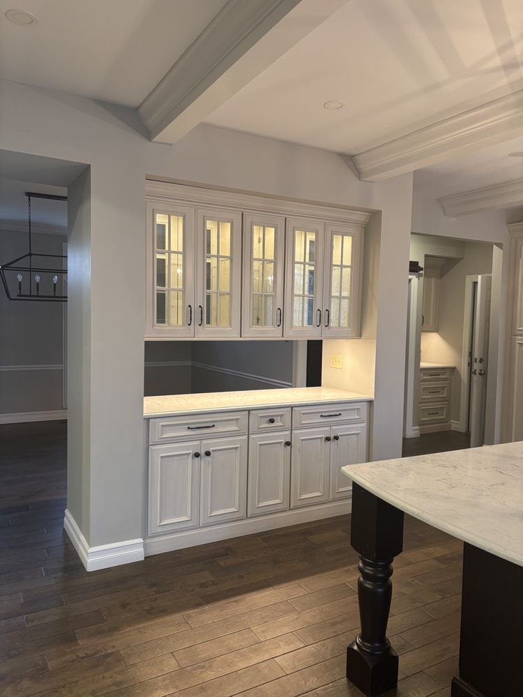 White built-in kitchen hutch with glass cabinets and under-cabinet lighting beside a marble island in a bright room