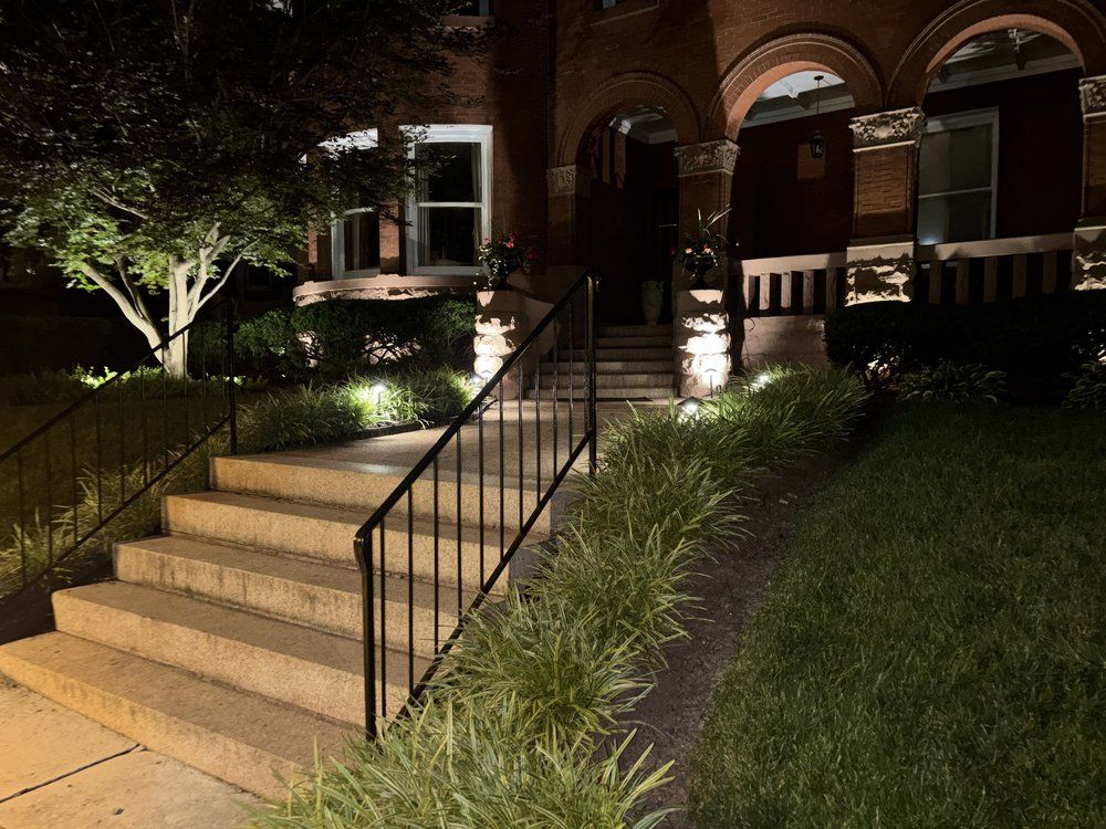 Lit front steps with black handrails leading to a brick building at night, surrounded by landscaping
