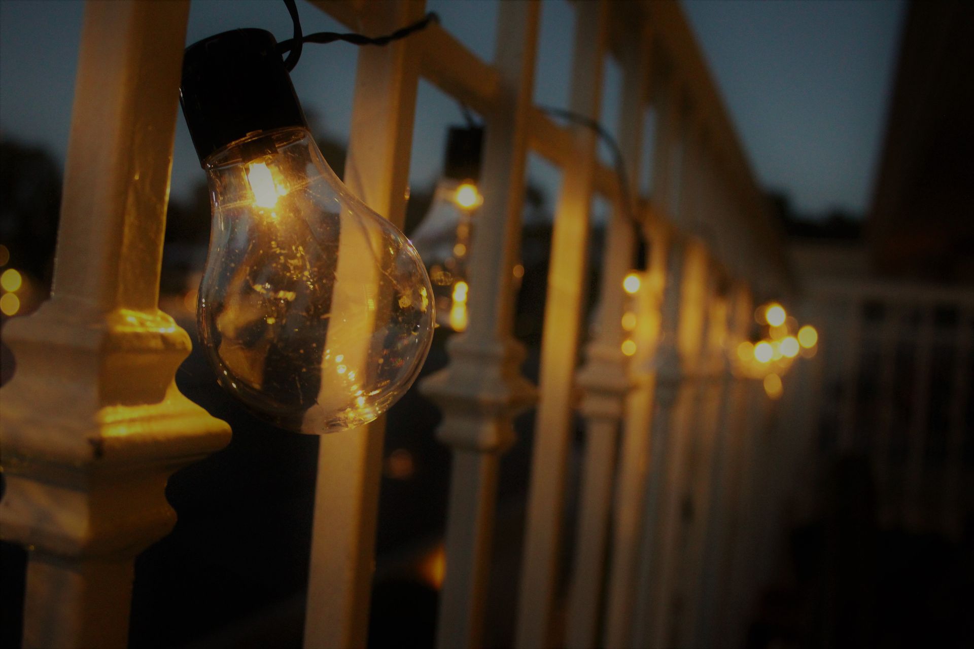 Warm string lights hanging along a wooden fence at dusk, with a glowing bulb in the foreground