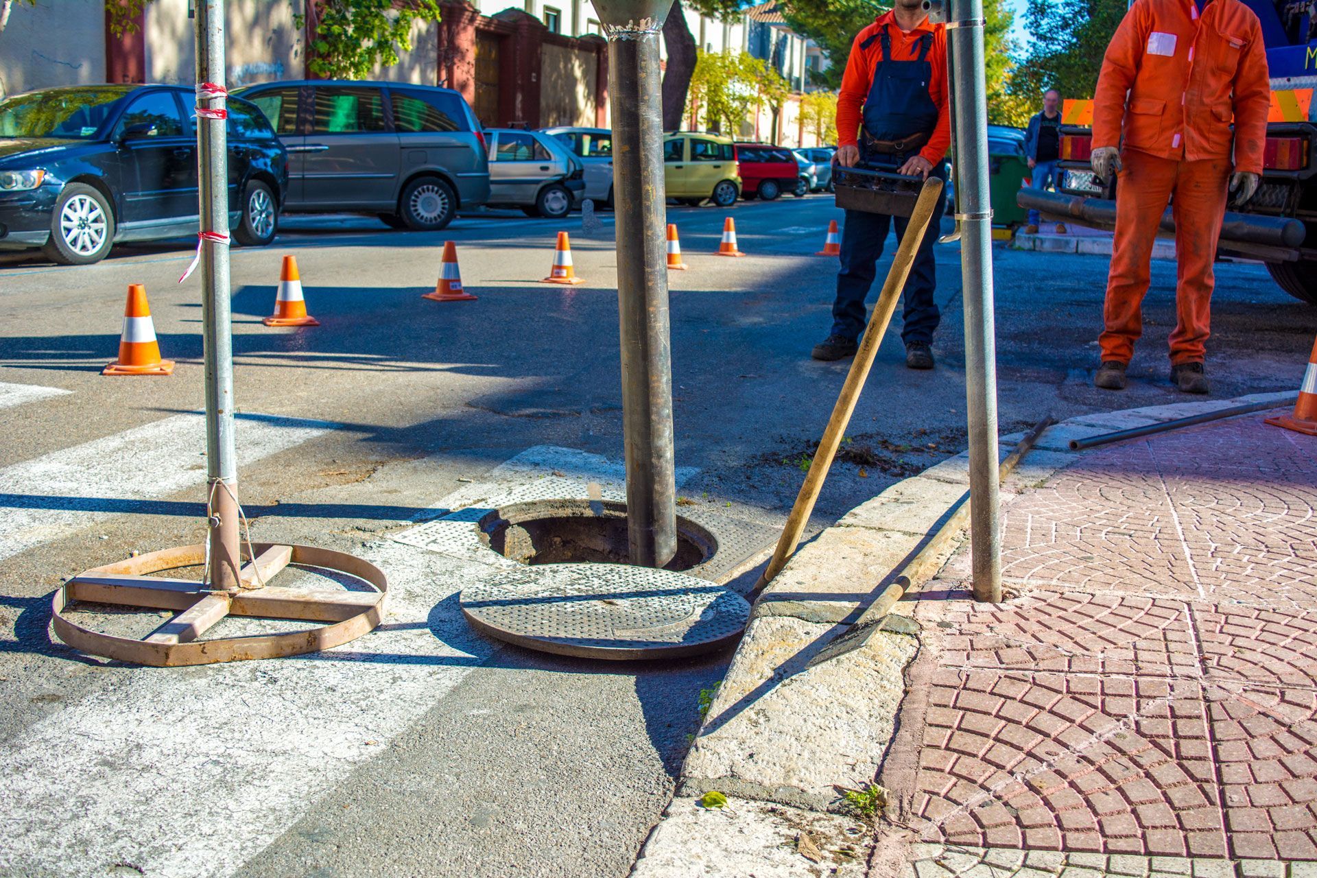 Road workers near an open manhole in a street, surrounded by safety cones. One worker holds a tool.