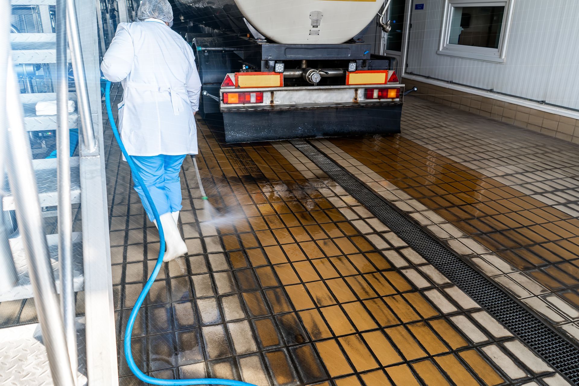Person in protective clothing sprays down a floor next to a tanker truck.