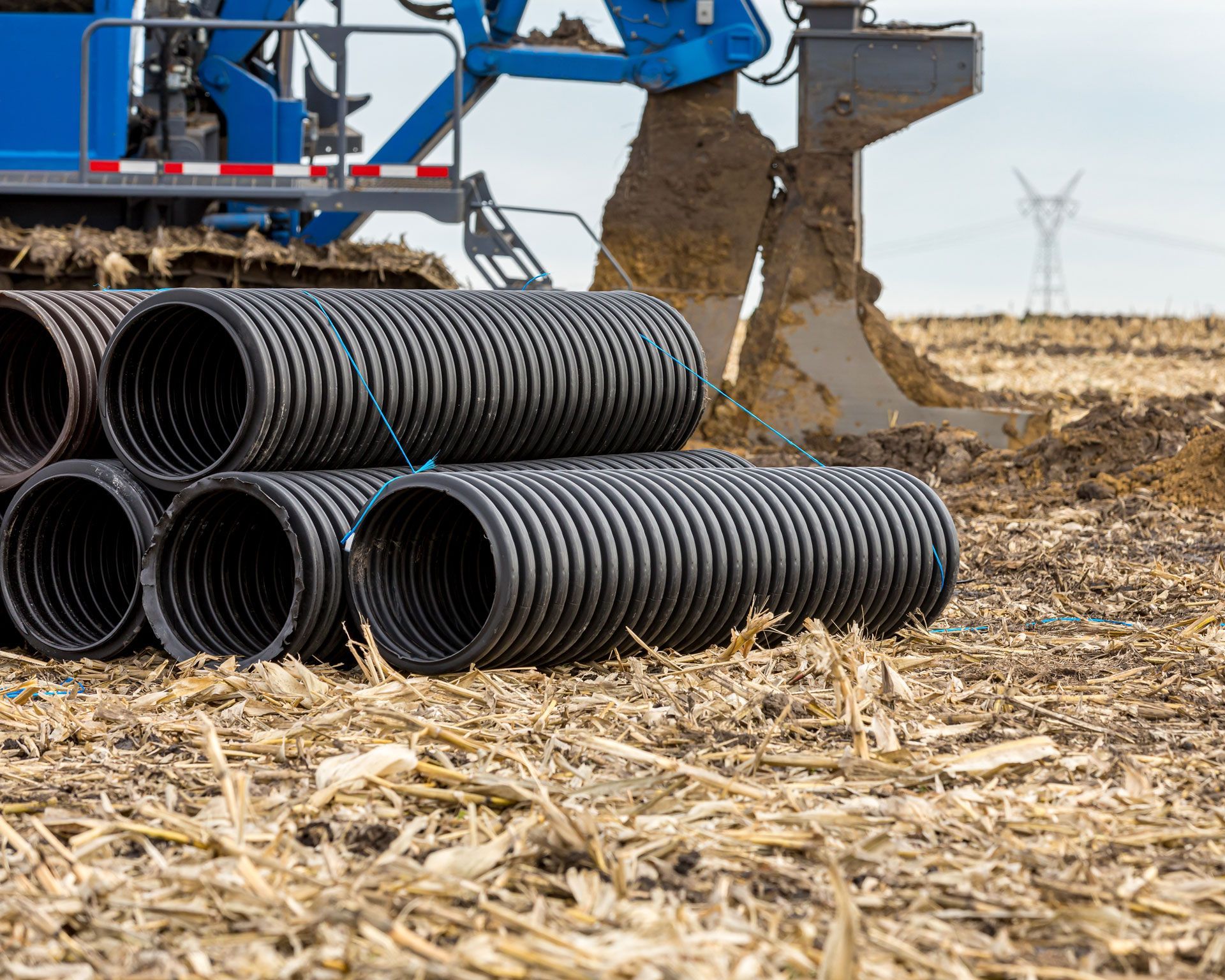 Black corrugated pipes next to construction equipment in a field, with power lines visible.