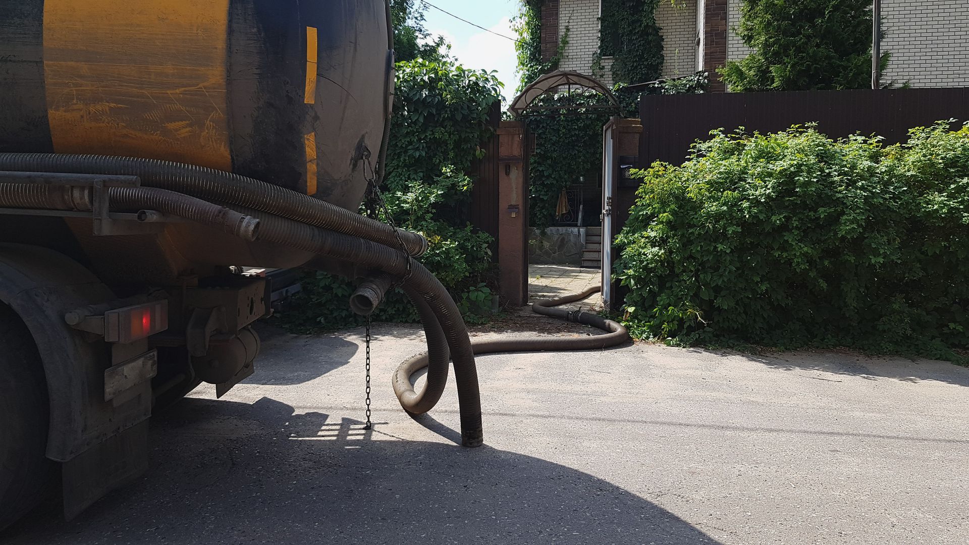 A large truck with a hose is parked near a gate with bushes; asphalt road.