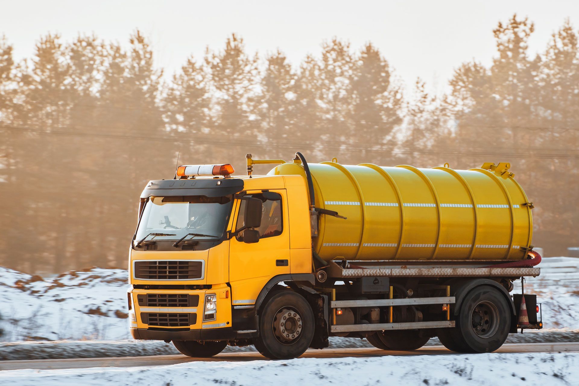 Yellow tanker truck driving on a snow-covered road. Trees in the background.