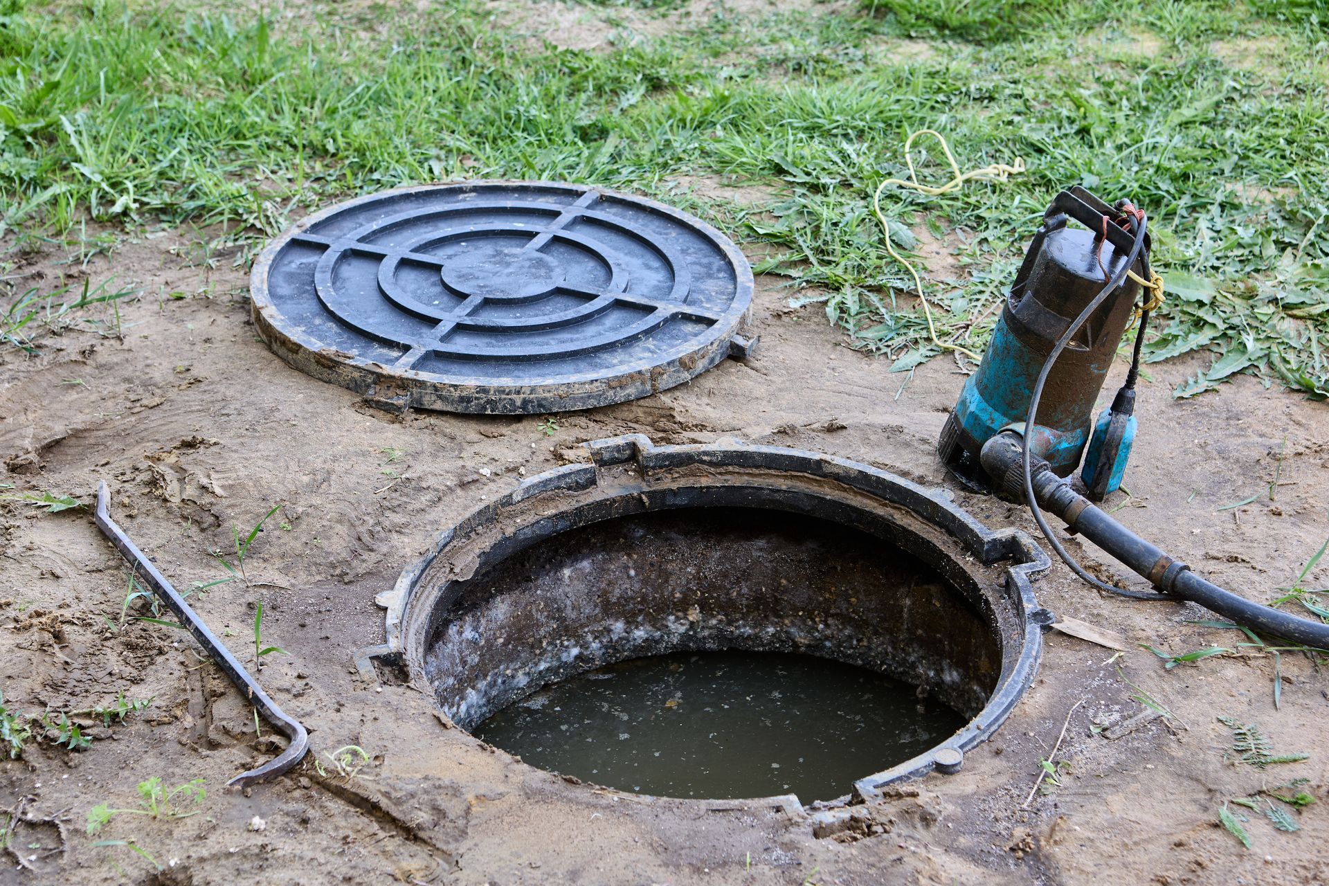Open manhole with water, detached cover, and a pump hose on a grassy dirt patch