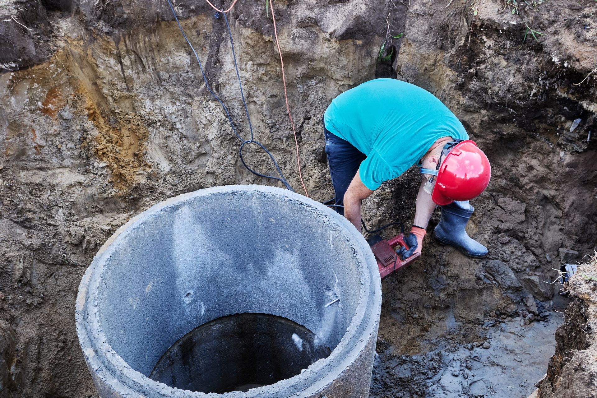 Worker wearing red hard hat cuts concrete pipe for septic system installation inside excavated pit.