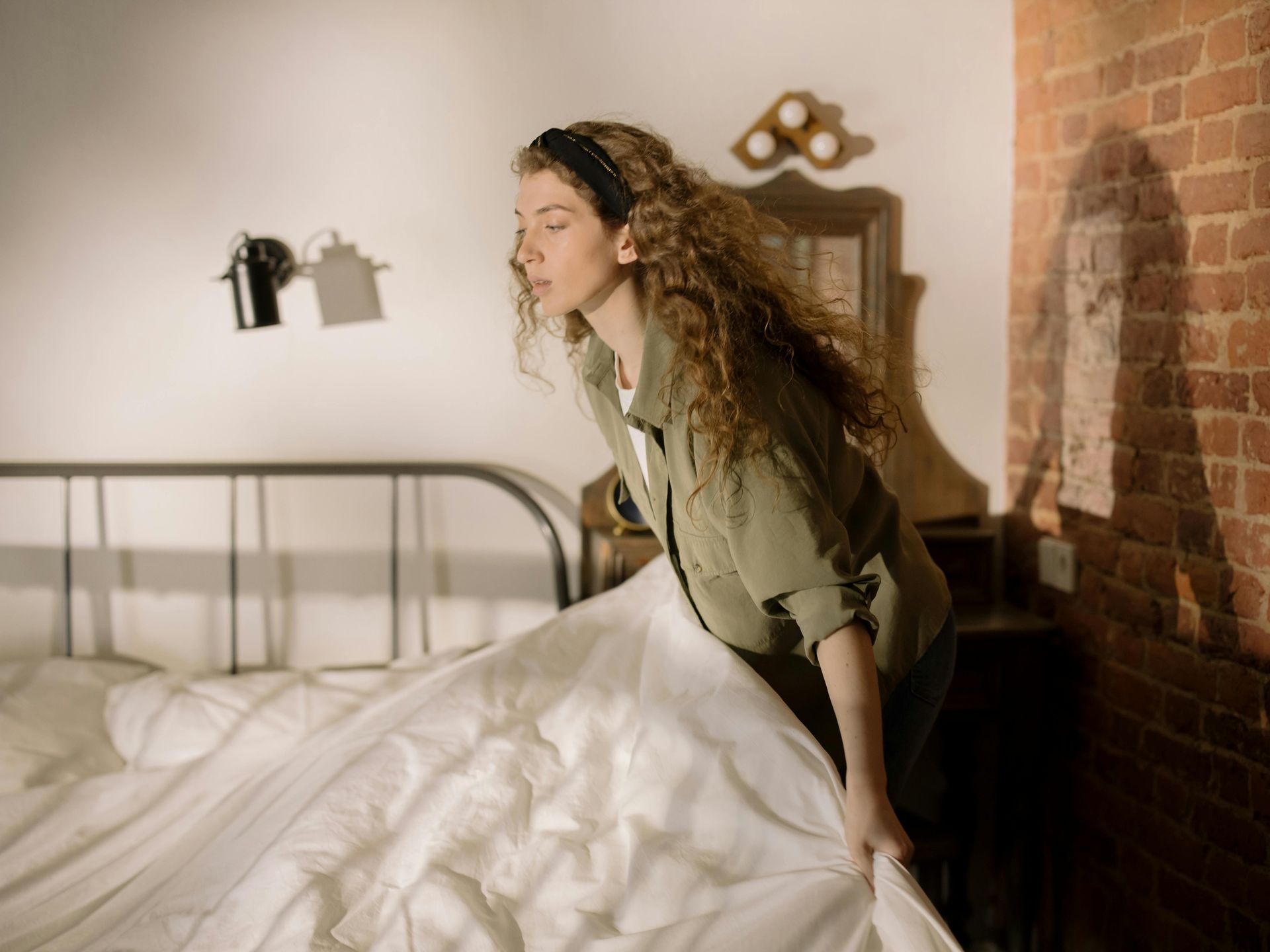 Een persoon met lang, golvend haar en een groen shirt staat naast een metalen bedframe en trekt een wit laken over het bed.