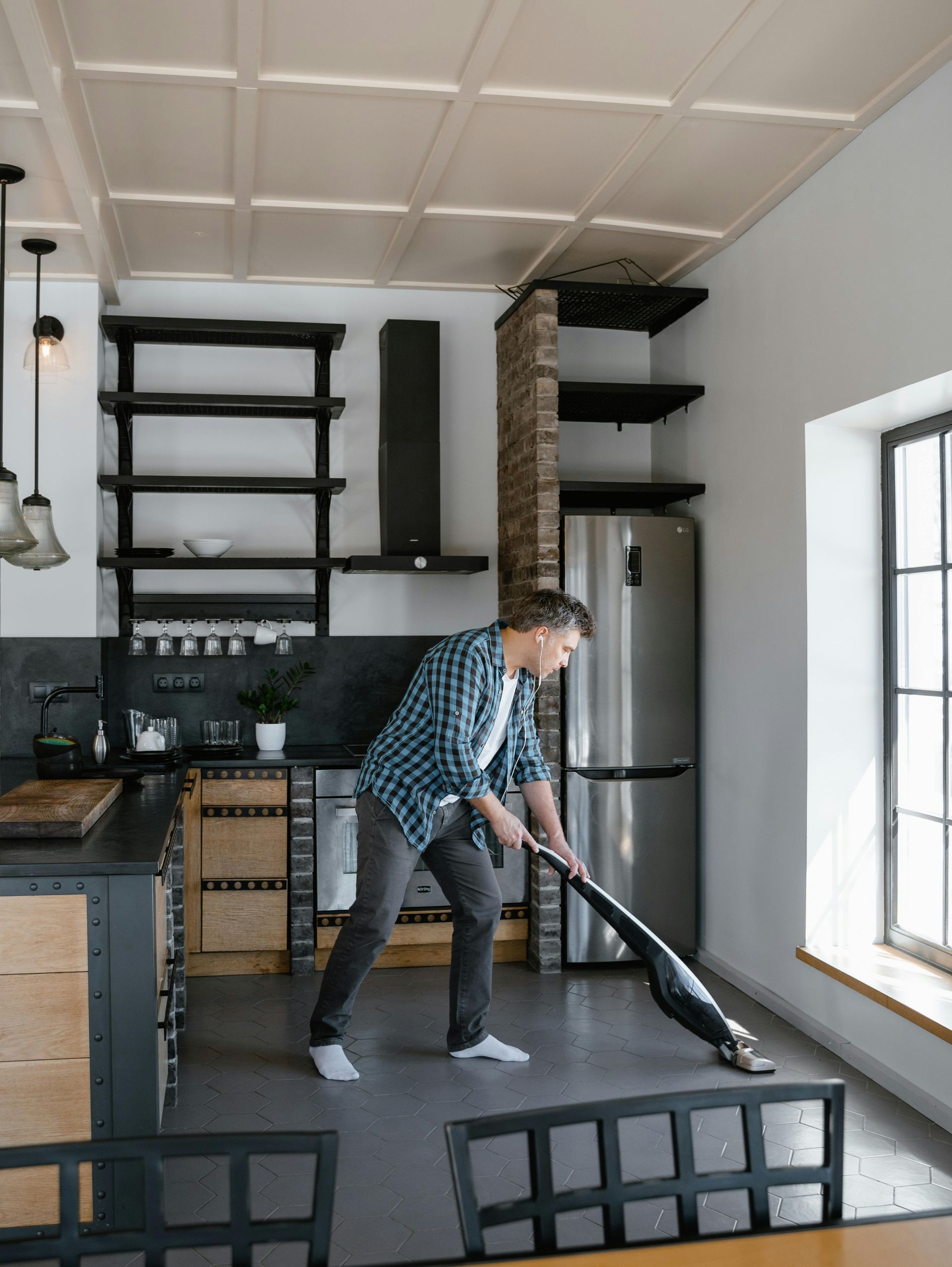 Een persoon in een geruit overhemd stofzuigt de vloer van een moderne keuken met zwarte planken, een bakstenen pilaar en roestvrij staal.