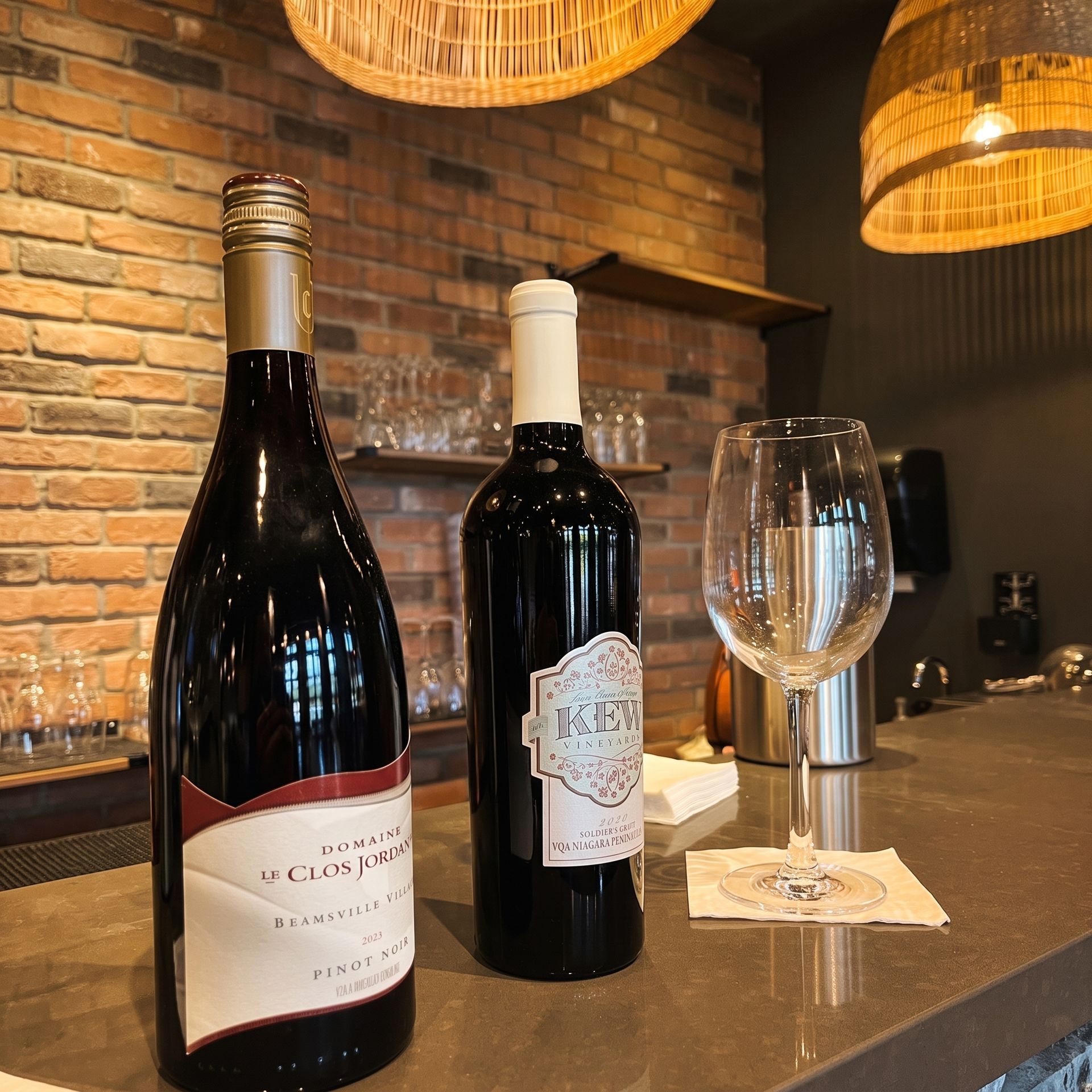 Two red wine bottles and a wine glass on a countertop in a brick-walled bar or restaurant