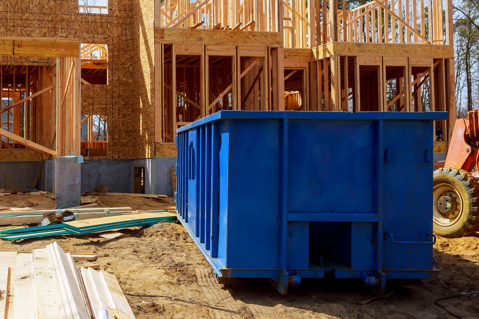 A large blue dumpster sits on the dirt ground in front of a house currently under construction.