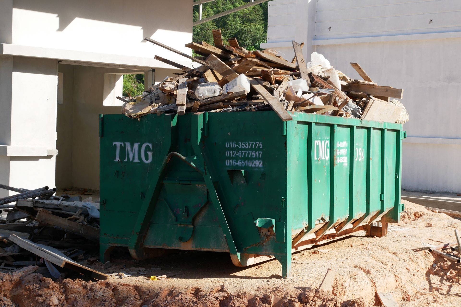 A large green TMG dumpster filled with construction debris sits on a dirt surface in front of a building.