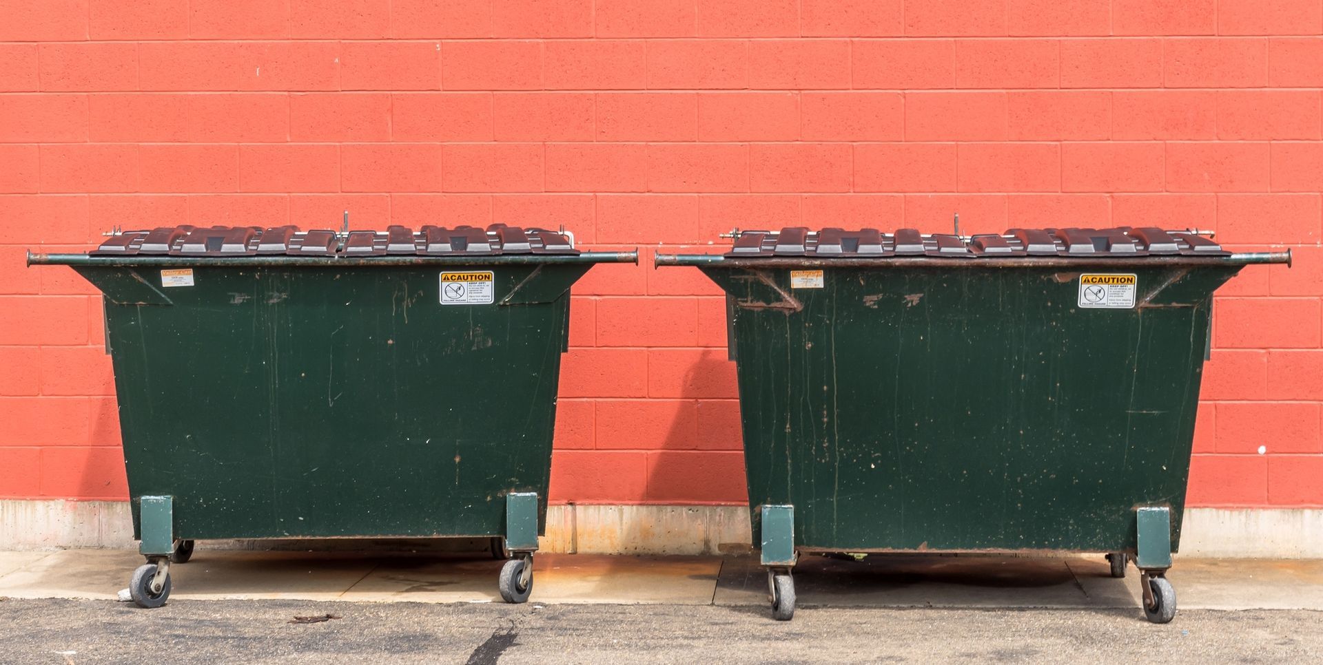 Two large dark green metal dumpsters sit side by side against a bright orange brick wall.
