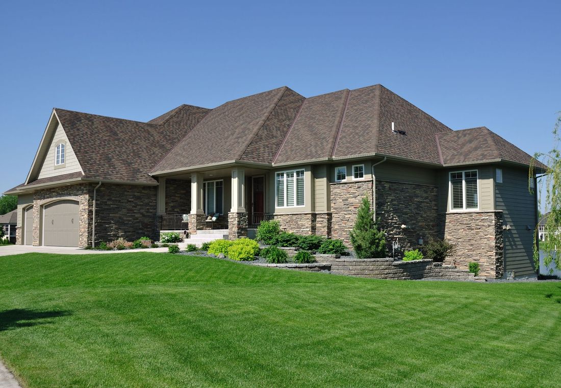 Large stone suburban house with a brown roof and green lawn under a clear blue sky