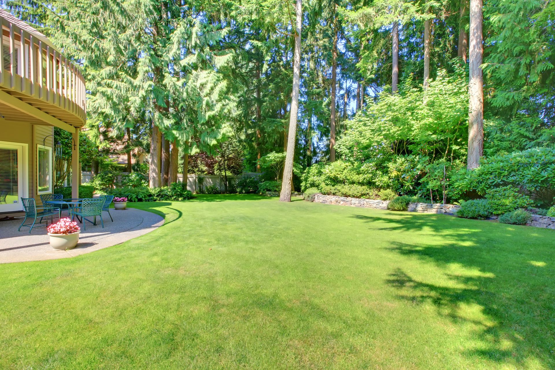 Lush green backyard lawn with tall trees, flowerbeds, and a house deck on the left.