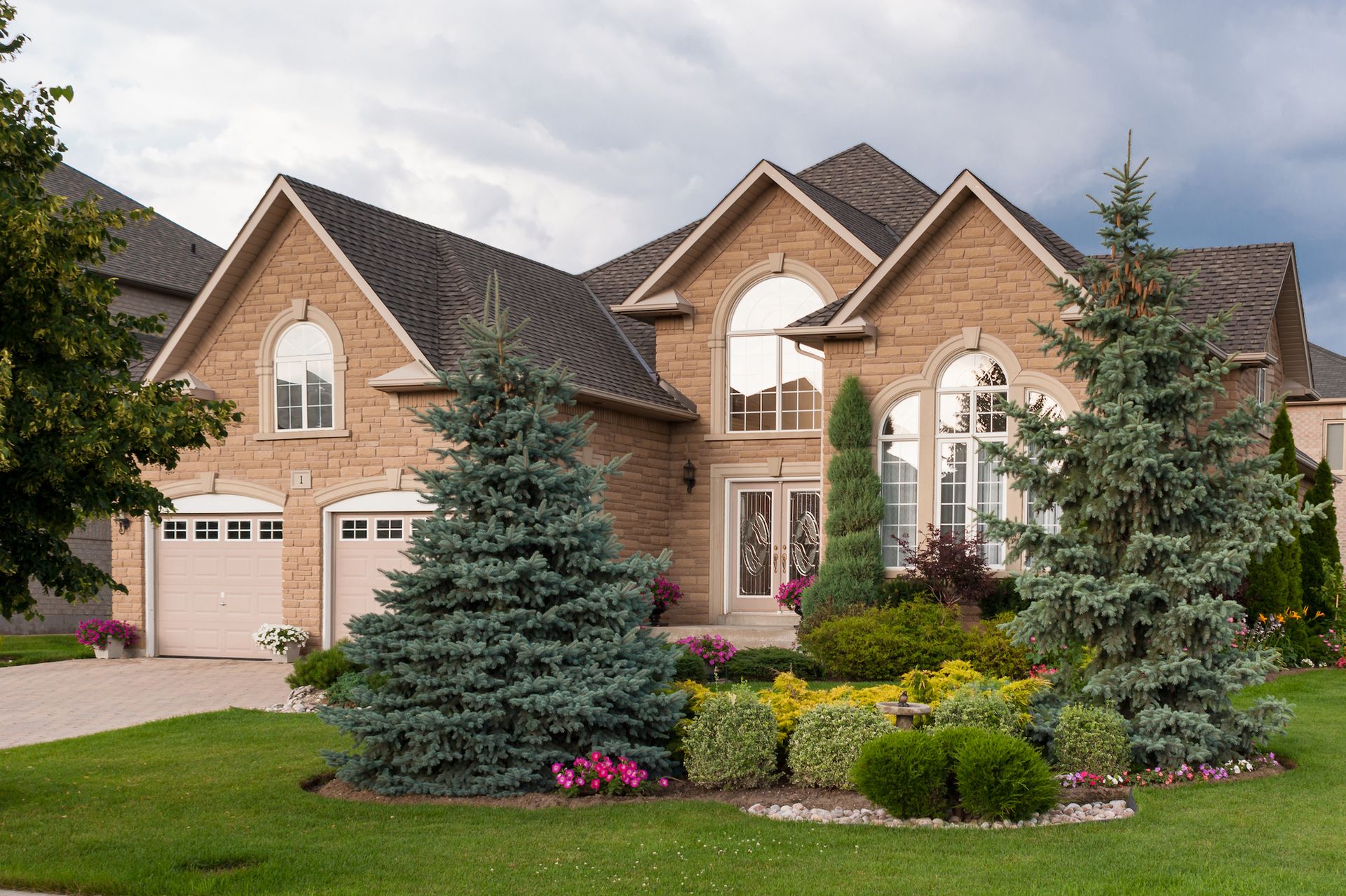 Large suburban brick house with manicured lawn, evergreen trees, and flower beds under a cloudy sky