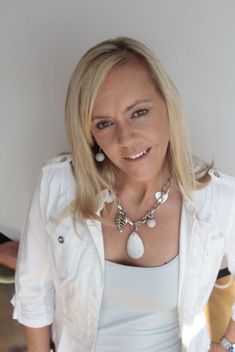 Blonde woman in white jacket and necklace smiling against a plain wall