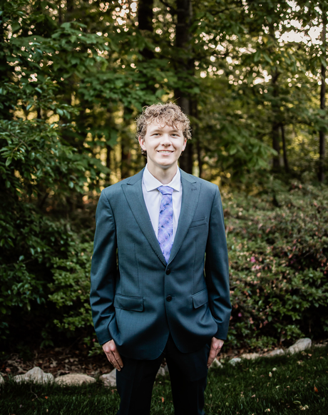 Person in a teal suit and lavender tie standing outdoors in a garden, smiling.