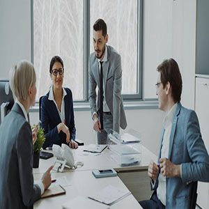 Business meeting around a conference table, with one man speaking and others listening in a bright office.