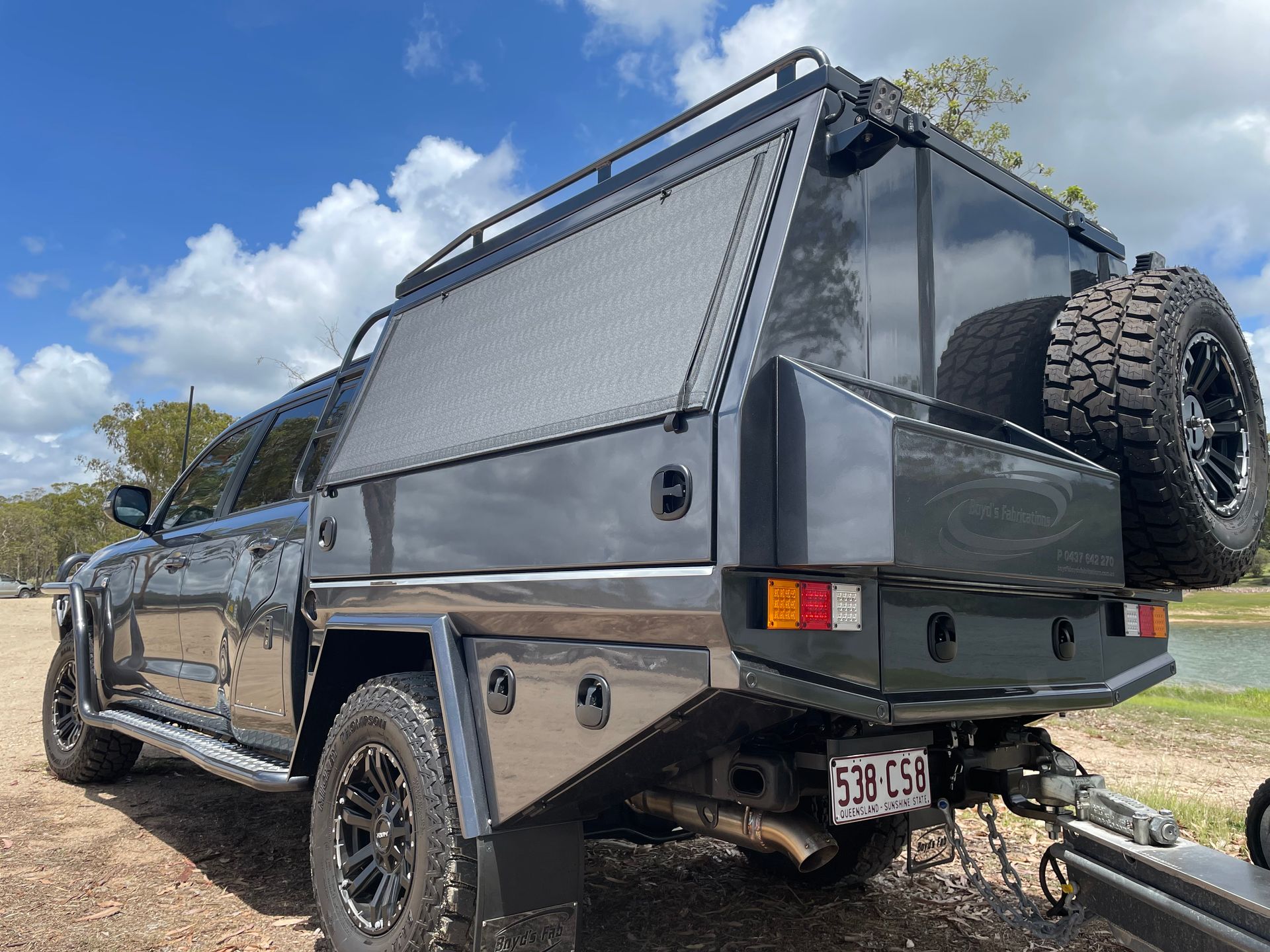 Dark gray pickup truck with a utility bed and spare tire parked near water on a sunny day.— Boyd's Fabrications In Portsmith, QLD 