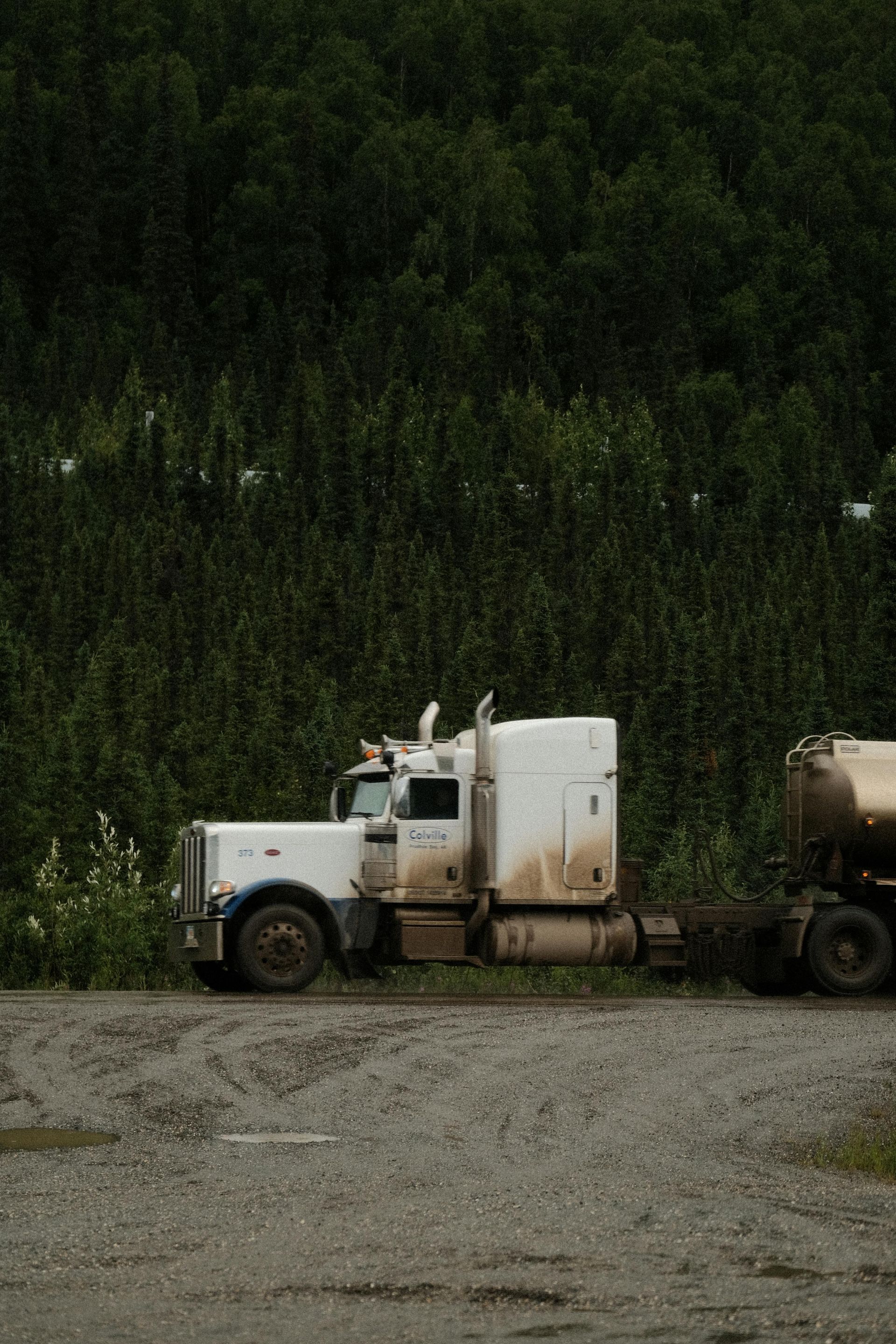 A semi truck is parked in a gravel lot in front of a forest.