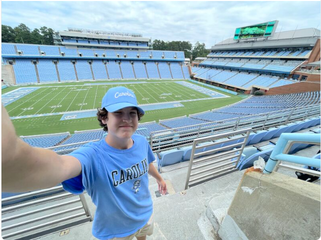 A man in a carolina shirt is taking a selfie in front of a football stadium