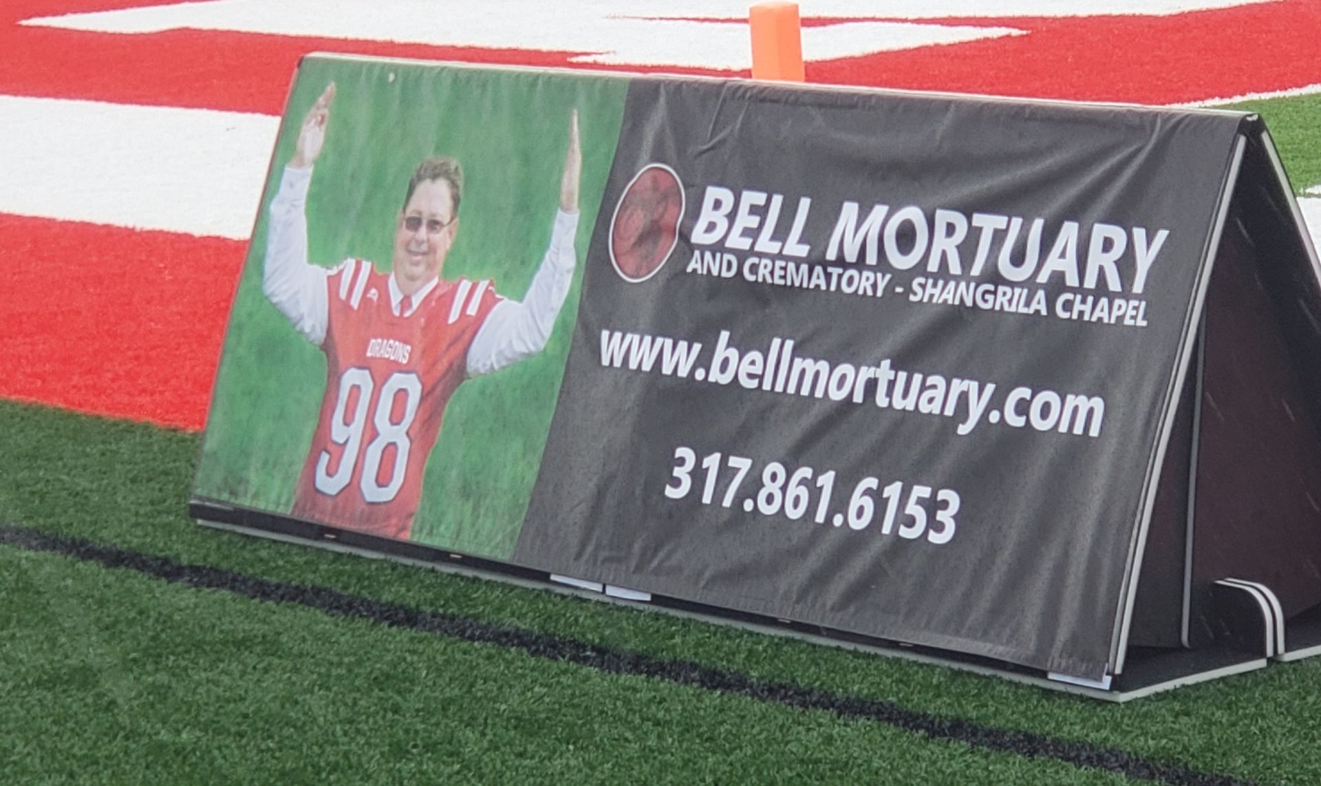 A banner for Bell Mortuary at a football game. It features a photo of a person and contact information.