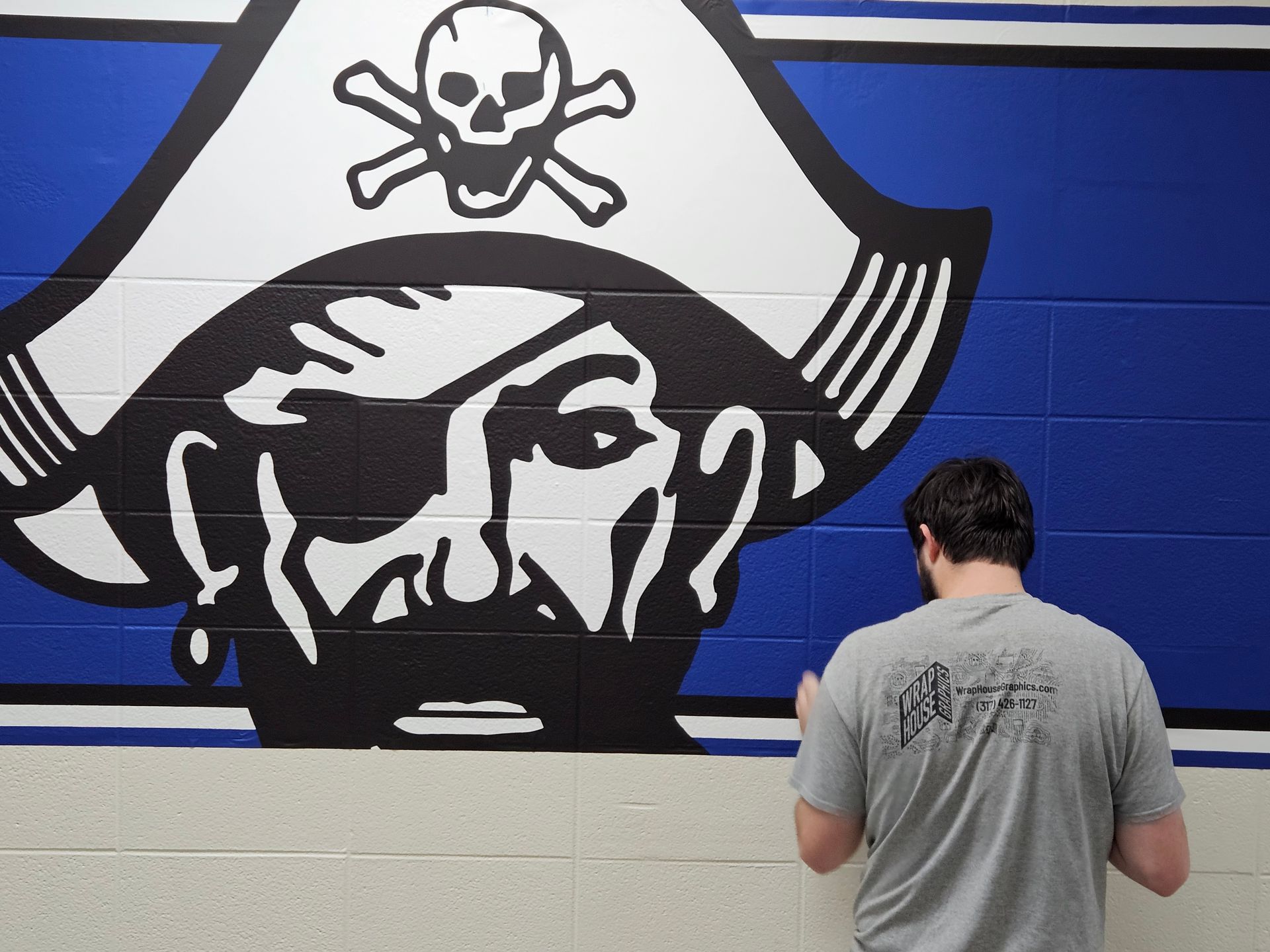 Man painting a large pirate logo on a blue and white wall.