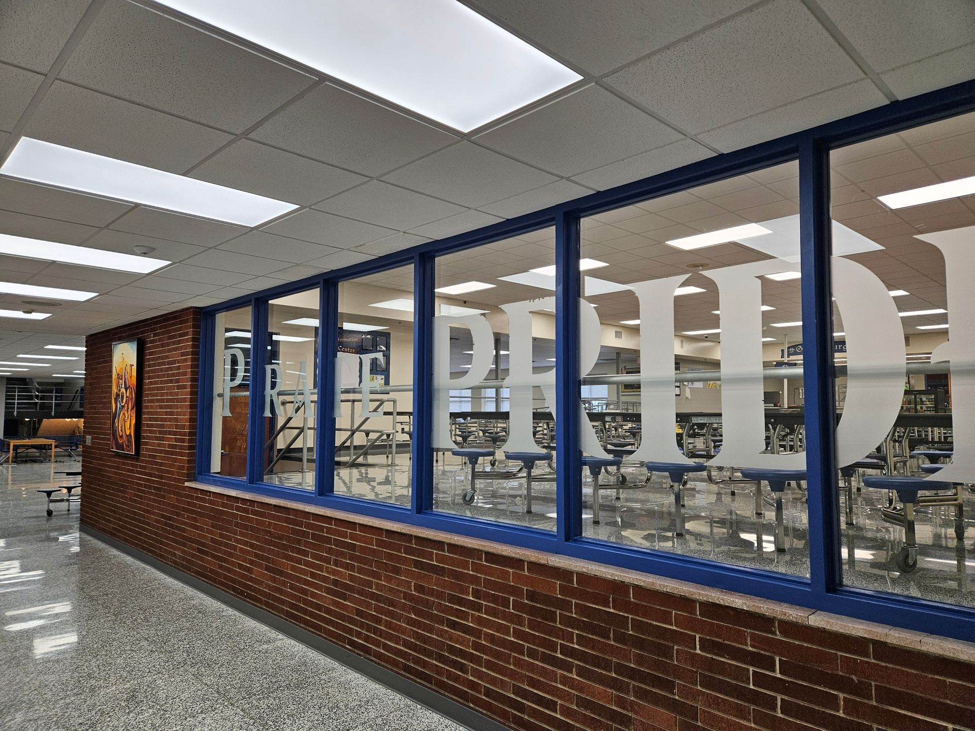Hallway with brick wall, blue-framed windows with "PHS" lettering, and school cafeteria visible through windows.