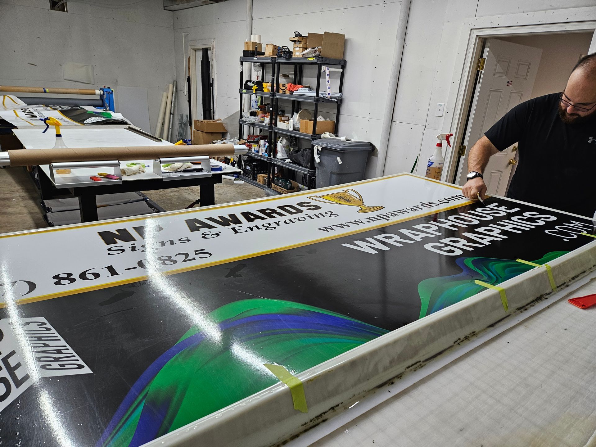 A man is applying graphics to a large sign in a workshop. The sign has green and blue swirls and text.