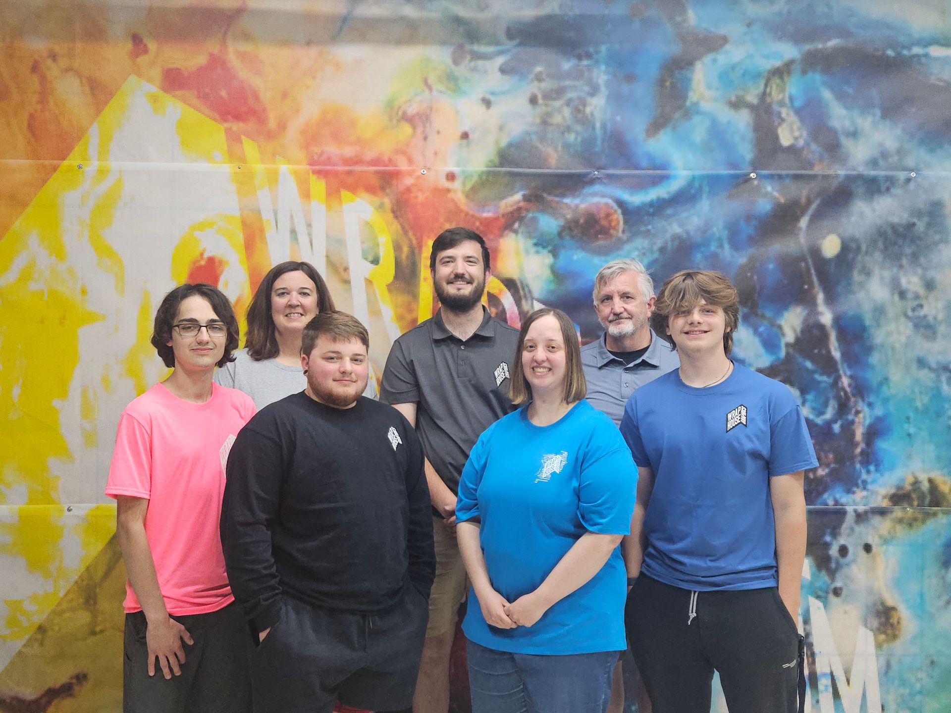Group of people in front of a colorful mural. People are wearing t-shirts and smiling.
