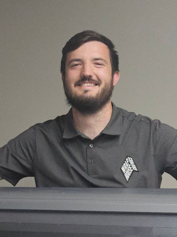 Man with beard smiling, wearing gray polo shirt with emblem. Arms outstretched, behind dark surface.