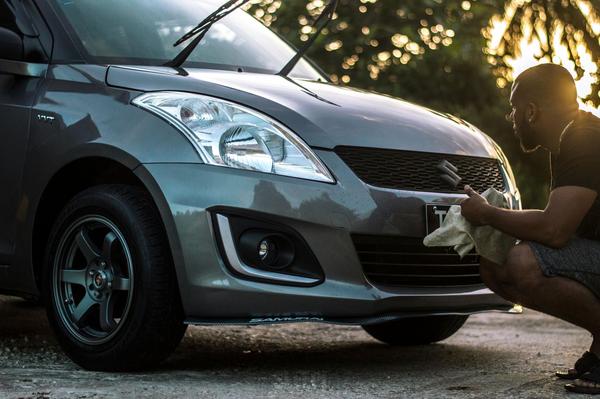 Man cleaning a gray car's front bumper, sunny outdoor setting.